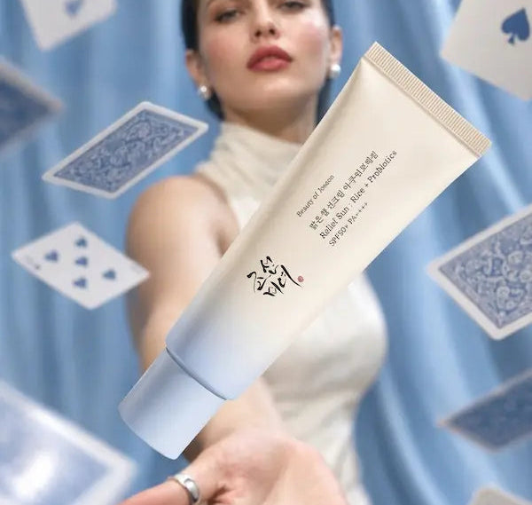 A woman in a white sleeveless dress extends her hand toward a floating beige cosmetic tube with Korean text, set against a blue draped backdrop with floating playing cards.