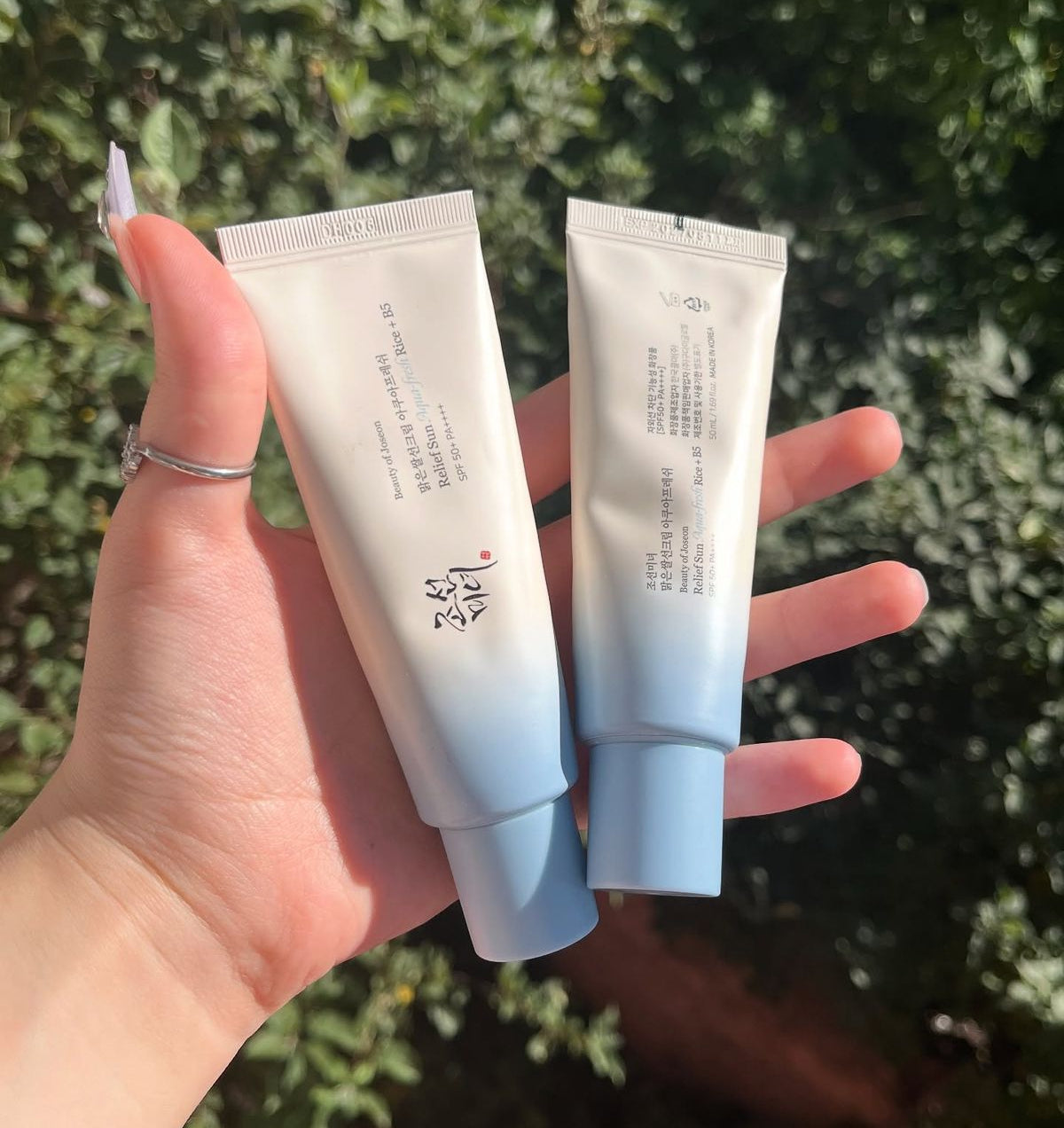 Two cosmetic squeeze tubes held in a person's hand outdoors, with a blurred green foliage background. The tubes are white at the top and fade to light blue at the bottom, with small black text and a small red logo mark.